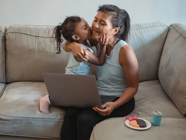 A mother working on a laptop at home while her daughter hugs her